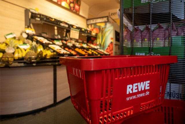 Ein Einkaufskorb mit dem Logo der Supermarktkette Rewe steht vor einer Obstauslage.