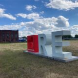 Auf der Brache des Gewerbegebiets Blumenthal in Recklinghausen steht auf einer verdorrten Wiese ein 3-D-Logo mit drei Elementen. Innerhalb eines roten Quaders ist ein Herz ausgespart worden, daneben stehen die Buchstaben R und E. Im Hintergrund ist ein altes Backstein-Industriegebäude zu erkennen.