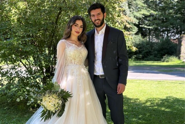 Eine Braut im weißen Hochzeitskleid und ein Mann im dunklen Anzug stehen im grünen Nahe beinander. Sie hält einen Strauß mit weißen Rosen in der Hand.