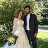 Eine Braut im weißen Hochzeitskleid und ein Mann im dunklen Anzug stehen im grünen Nahe beinander. Sie hält einen Strauß mit weißen Rosen in der Hand.