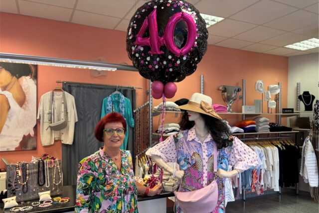 Eine Frau in einem Geschäft trägt ein buntes Kleid. In der Hand hält sie einen Ballon, auf dem eine große 40 steht. Neben ihr steht eine Schaufensterpuppe, auch farbenfroh gekleidet.