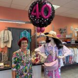 Eine Frau in einem Geschäft trägt ein buntes Kleid. In der Hand hält sie einen Ballon, auf dem eine große 40 steht. Neben ihr steht eine Schaufensterpuppe, auch farbenfroh gekleidet.
