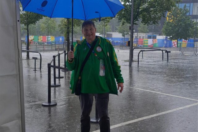 EM 2024-Volunteer Peter Planke steht mit einem blauen Regenschirm im strömenden Regen vor dem Dortmunder Stadion.