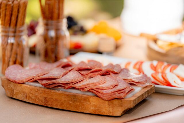 Geschnittene Salami-Scheiben liegen auf einem Holzbrett, das mit weiteren Lebensmitteln auf einem Tisch steht.