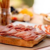 Geschnittene Salami-Scheiben liegen auf einem Holzbrett, das mit weiteren Lebensmitteln auf einem Tisch steht.