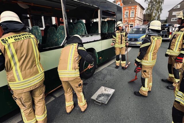 Feuerwehrleute stehen neben einem Bus auf einer Straße. Seitenscheiben des Busses sind zersplittert. Im Hintergrund sind Häuser und Einsatzfahrzeuge der Polizei zu sehen.
