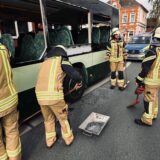 Feuerwehrleute stehen neben einem Bus auf einer Straße. Seitenscheiben des Busses sind zersplittert. Im Hintergrund sind Häuser und Einsatzfahrzeuge der Polizei zu sehen.