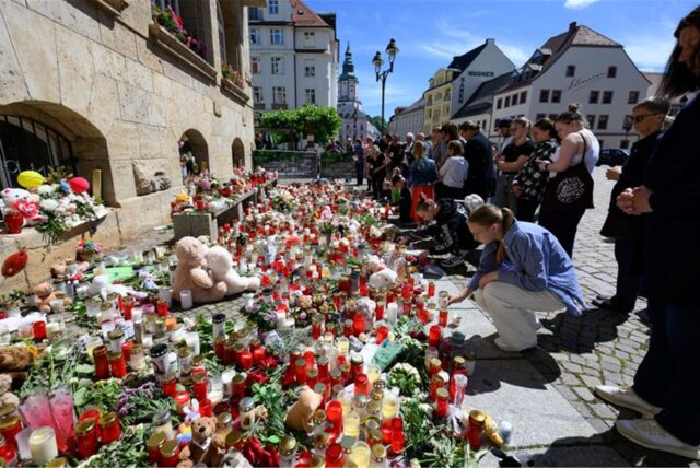 Menschen stehen am Rande eines Gottesdienstes für die getötete Valeriia in Döbeln vor Blumen und Kerzen.