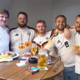 Fünf Männer in Deutschland-Trikot stehen an einem Tisch mit Getränken und etwas zu essen.