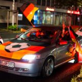 Autokorso in Recklinghausen: In einem vor einer roten Ampel auf dem Wallring stehenden Opel sitzen Fans der deutschen Fußballnationalmannschaft und schwenken Flaggen. Über die Motorhaube des Autos ist eine schwarz-rot-goldene Flagge mit einem Fußball in der Mitte gespannt.