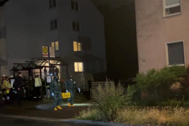 Zwei Spezialeinsatzkräfte tragen in Schutzanzügen und unter Atemschutz eine Kiste aus einem Gebäude an der Hochstraße.