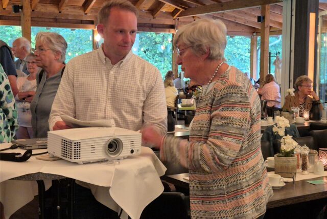 Ein Mann und eine Frau stehen an einem Stehtisch und unterhalten sich. Im Hintergrund sitzen Menschen an gedeckten Tischen. Auf dem Stehtisch steht ein Beamer.