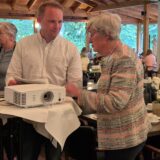 Ein Mann und eine Frau stehen an einem Stehtisch und unterhalten sich. Im Hintergrund sitzen Menschen an gedeckten Tischen. Auf dem Stehtisch steht ein Beamer.