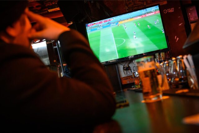 Ein Mann sitzt an der Bar in und schaut auf einem Fernseher Fußball.
