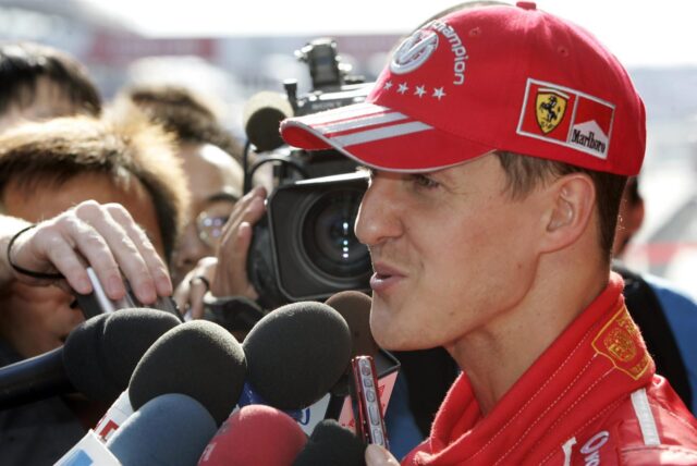 Ex-Rennfahrer Michael Schumacher spricht während eines Interviews in mehrere Mikrofone. Er trägt eine rote Jacke und eine rote Kappe.