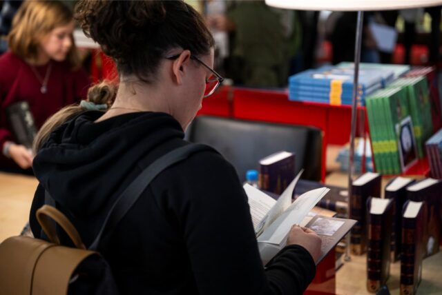 Eine Frau steht vor einem Tisch, auf dem mehrere Bücher ausgelegt sind. Sie selbst hält eines in den Händen und blättert es durch.