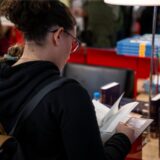 Eine Frau steht vor einem Tisch, auf dem mehrere Bücher ausgelegt sind. Sie selbst hält eines in den Händen und blättert es durch.
