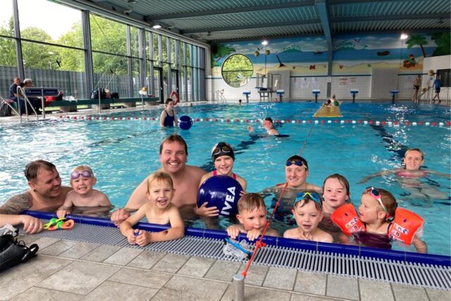 Zehn Kinder und Erwachsene in Schwimmkleidung lehnen sich in einem Schwimmbecken an den Beckenrand und lächeln in die Kamera.