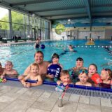 Zehn Kinder und Erwachsene in Schwimmkleidung lehnen sich in einem Schwimmbecken an den Beckenrand und lächeln in die Kamera.