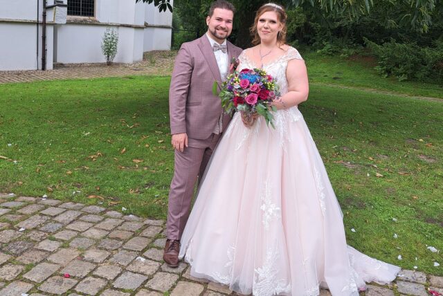 Braut mit Blumenstrauß in zartes Rosa gekleidet und Bräutigam im dazu passenden hellen Anzug, lächeln in die Kamera. Im Hintergrund ist es grün und ein Teil der weißen Schlosskapelle ist zu sehen.