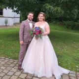 Braut mit Blumenstrauß in zartes Rosa gekleidet und Bräutigam im dazu passenden hellen Anzug, lächeln in die Kamera. Im Hintergrund ist es grün und ein Teil der weißen Schlosskapelle ist zu sehen.