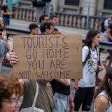 Eine Gruppe von Menschen protestiert gegen die negativen Effekte des Tourismus in Barcelona. Ein Mann hält ein Plakat hoch auf dem steht „Tourists go home, you are not welcome“.