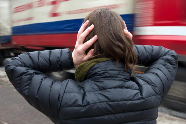 Eine Frau hält sich die Ohren zu, ein Schwerlaster fährt vorbei.