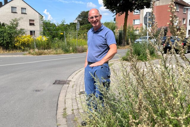 Ein Mann steht mit Jeans und blauem Polohemd an einem Straßenrand in Recklinghausen und zeigt mit einer Handbewegung den hochgewachsenen Grünstreifen.
