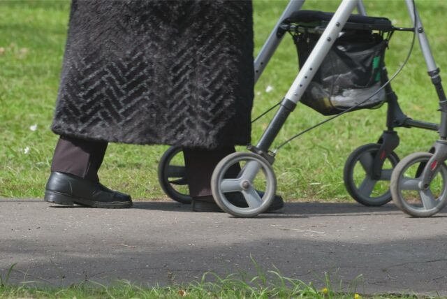 Eine Frau in schwarzem Mantel, die nur bis zu den Knien zu sehen ist, geht mit einem Rollator über einen asphaliterten Weg. (Symbolbild)