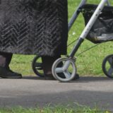 Eine Frau in schwarzem Mantel, die nur bis zu den Knien zu sehen ist, geht mit einem Rollator über einen asphaliterten Weg. (Symbolbild)