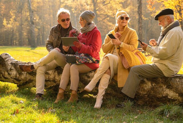 Befreundete Senioren sitzen im Herbst mit ihren mobilen Endgeräten im Park.