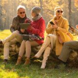 Befreundete Senioren sitzen im Herbst mit ihren mobilen Endgeräten im Park.