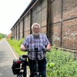 Ein Mann steht mit kariertem Hemd und einem Fahrrad auf einem Radweg. Hinter ihm ist ein historisches Gebäude aus dem Bergbau zu sehen.