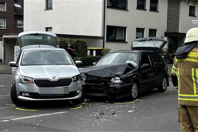 Zwei Autos stehen nach einem Unfall auf einer Straße. Rechts ist ein Feuerwehrmann zu sehen. Ein schwarzes Auto hat schweren Schaden davongetragen.