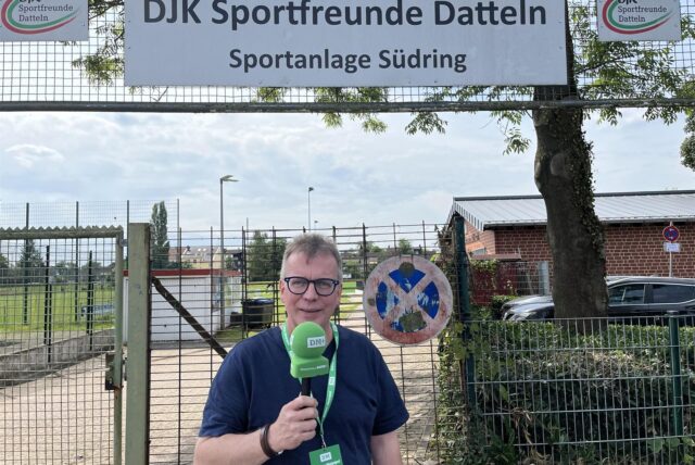 Redakteur Uwe Wallkötter steht vor einer Sportanlage in Datteln. Er hält ein Mikrofon in der Hand.