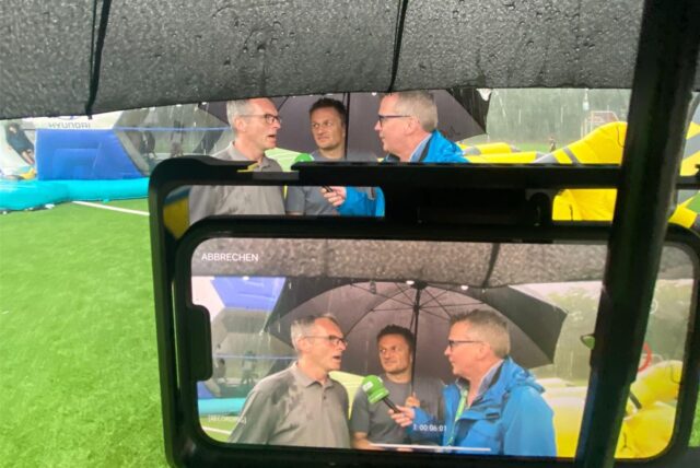 Drei Männer stehen während eines Interviews auf einem Sportplatz unter einem Regenschirm, im Vordergrund eine Handykamera, die die Szene filmt.