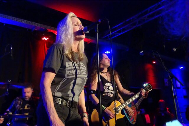 Eine Frau mit einem schwarzen T-Shirt steht auf einer Bühne und singt in ein Mikrofon.