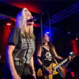 Eine Frau mit einem schwarzen T-Shirt steht auf einer Bühne und singt in ein Mikrofon.