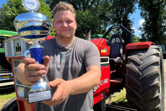 Ein Mann vor einem Traktor hält einen Pokal in die Kamera.