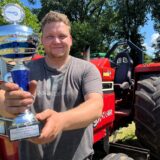 Ein Mann vor einem Traktor hält einen Pokal in die Kamera.
