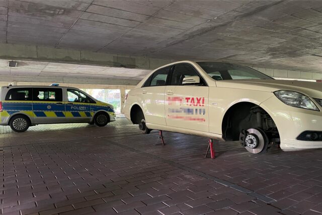 Ein Taxi steht aufgebockt und ohne Reifen in der unteren Etage eines Parkhauses. Im Hintergrund steht ein Polizeiwagen.