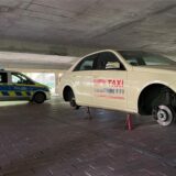 Ein Taxi steht aufgebockt und ohne Reifen in der unteren Etage eines Parkhauses. Im Hintergrund steht ein Polizeiwagen.