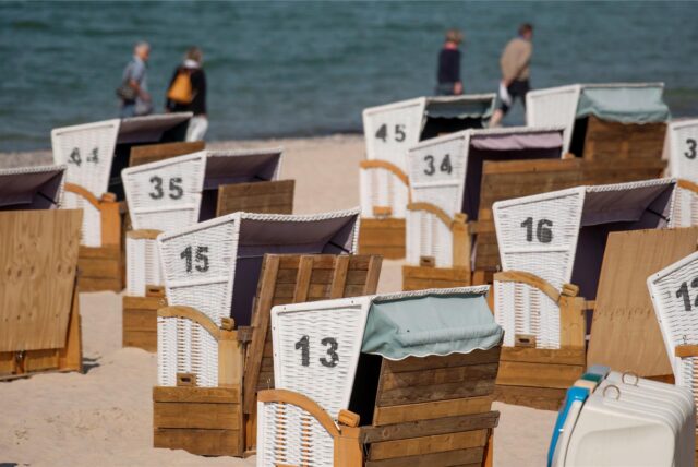 Menschen gehen am Samstag bei warmem, sonnigen Wetter an verschlossenen Strandkörben am Ostseestrand vorbei.