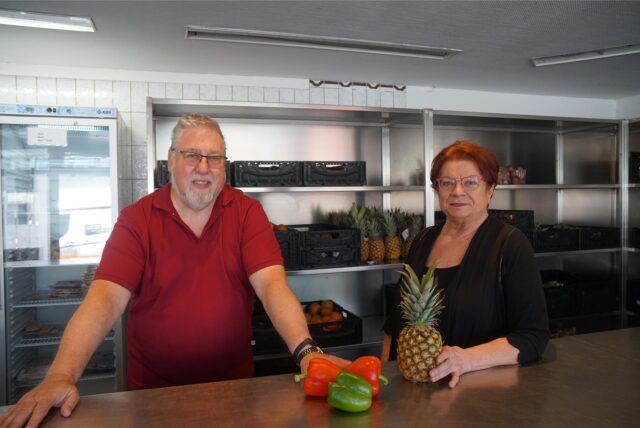 Wolfgang Schulz und Edeltraud Hartelt stehen in den Räumen der Marler Tafel. Hinter ihnen stehen Regale voller Lebensmittel. Auf dem Tisch vor ihnen liegen drei Paprika und eine Ananas.