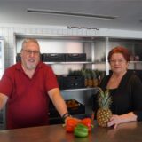 Wolfgang Schulz und Edeltraud Hartelt stehen in den Räumen der Marler Tafel. Hinter ihnen stehen Regale voller Lebensmittel. Auf dem Tisch vor ihnen liegen drei Paprika und eine Ananas.