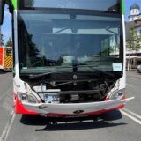 Ein Bus mit leicht demolierter Front steht an einem heißen Sommertag auf einer Straße. Im Hintergrund sind Rettungswagen zu sehen.