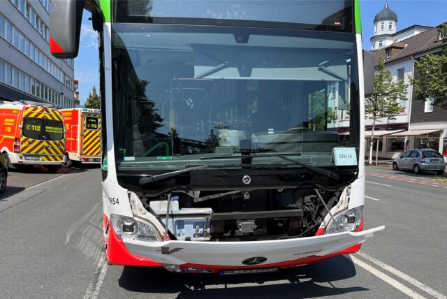 Ein Bus mit leicht demolierter Front steht an einem heißen Sommertag auf einer Straße. Im Hintergrund sind Rettungswagen zu sehen.