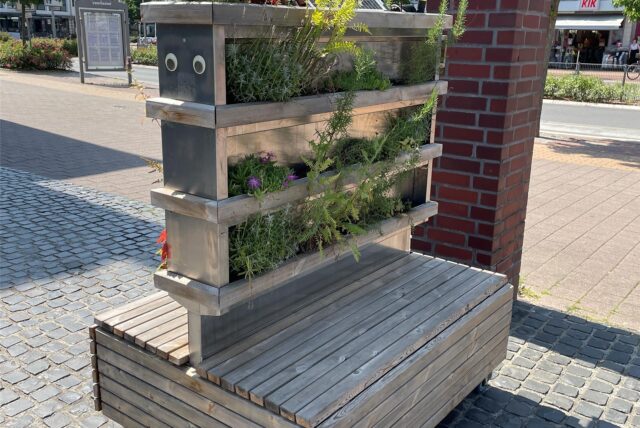 Ein vertikaler Blumenkasten aus Holzlatten und Metallelementen steht auf einer gepflasterten Fläche in Datteln. Im Hintergrund eine geklinkerte Säule und die Fahrbahn einer Straße,