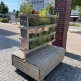 Ein vertikaler Blumenkasten aus Holzlatten und Metallelementen steht auf einer gepflasterten Fläche in Datteln. Im Hintergrund eine geklinkerte Säule und die Fahrbahn einer Straße,