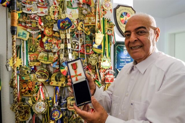 Mehdi Noheh-Khan aus Recklinghausen steht lächelnd mit weißem Hemd vor einer Wand voller Medaillen und Orden. In der Hand hält er eine Schatulle mit einem Bundesverdienstkreuz.